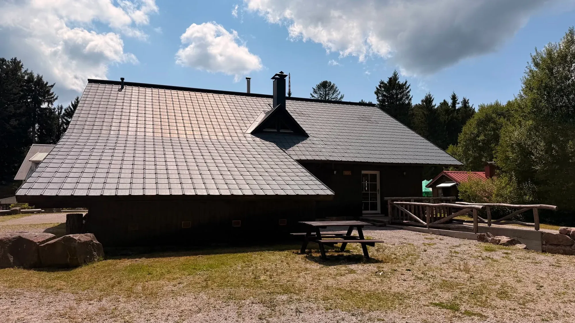Seitenansicht der Skihütte mit Sitzbank, Tischen und Bäumen unter blauem Himmel.