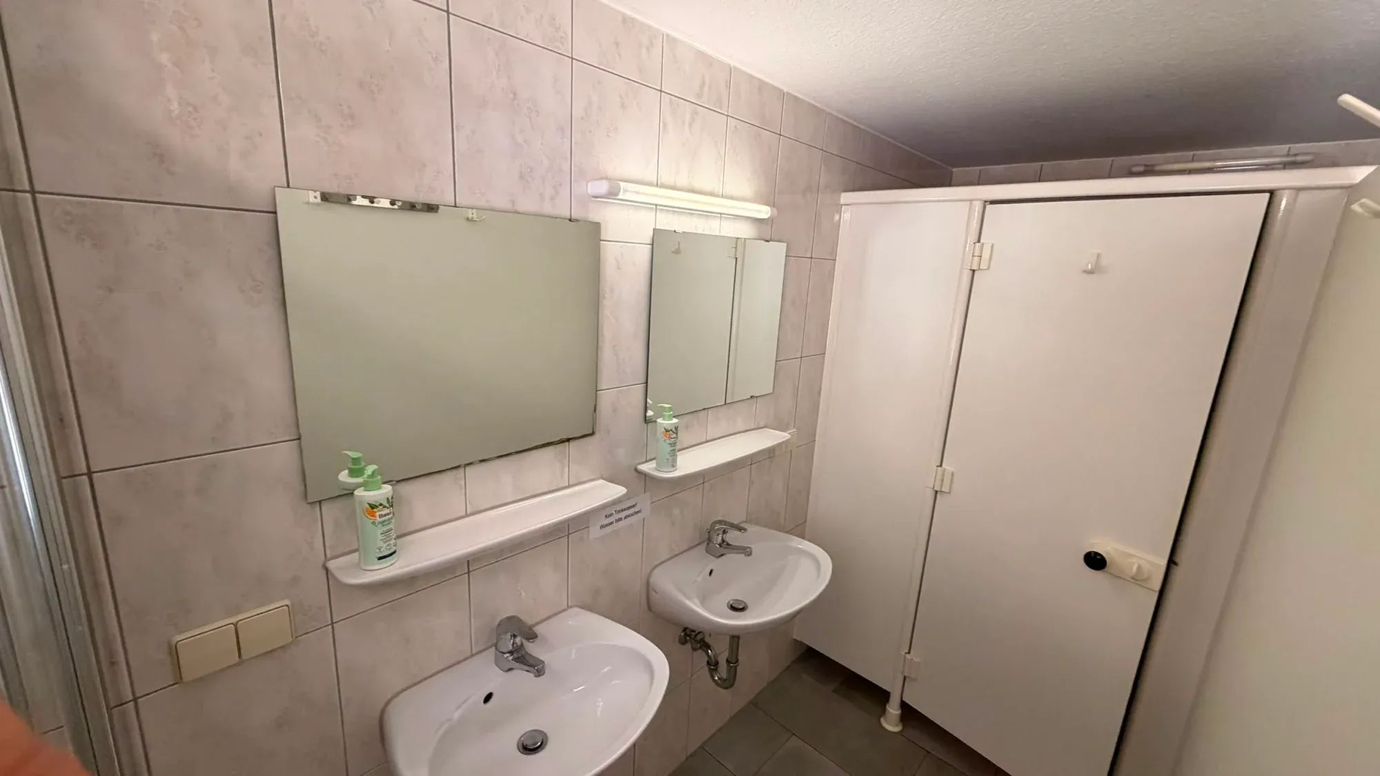 Gefliester Waschraum mit zwei Waschbecken, Spiegeln und Duschkabinen.